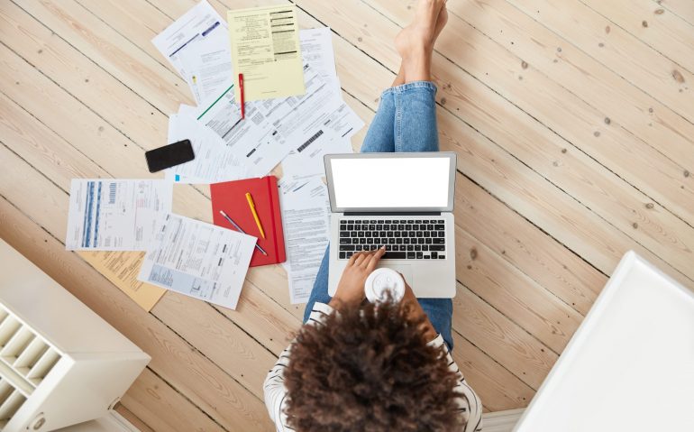 Top view of darrk skinned female deals with bills and taxes, works on laptop computer with white mock up screen, drinks takeaway coffe, poses on wooden floor at domestic setting, makes report